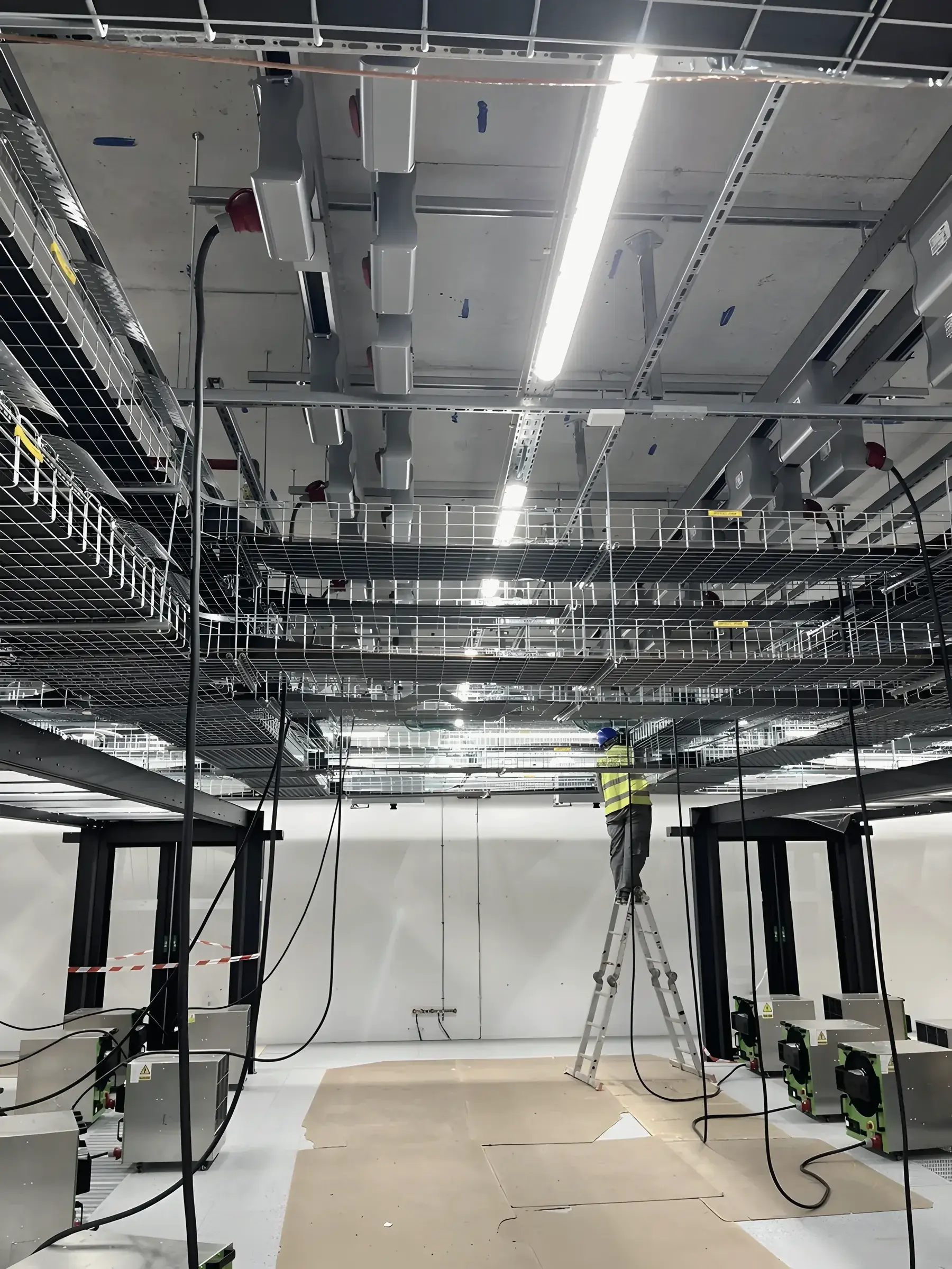 A technician on a ladder fixing cable tray under the ceiling of a data hall during fit-out