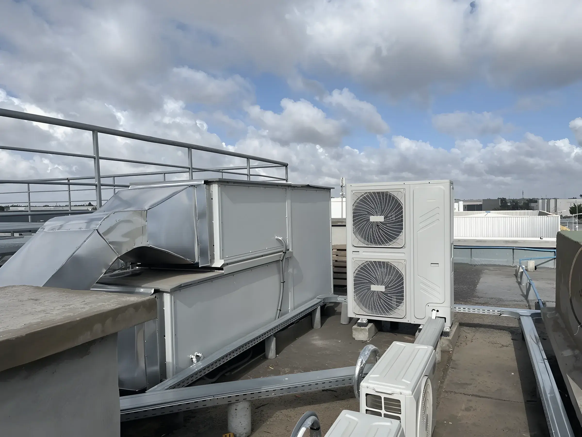 A galvanised duct and a condensing unit on the roof, with a railing and a cloud sky behind