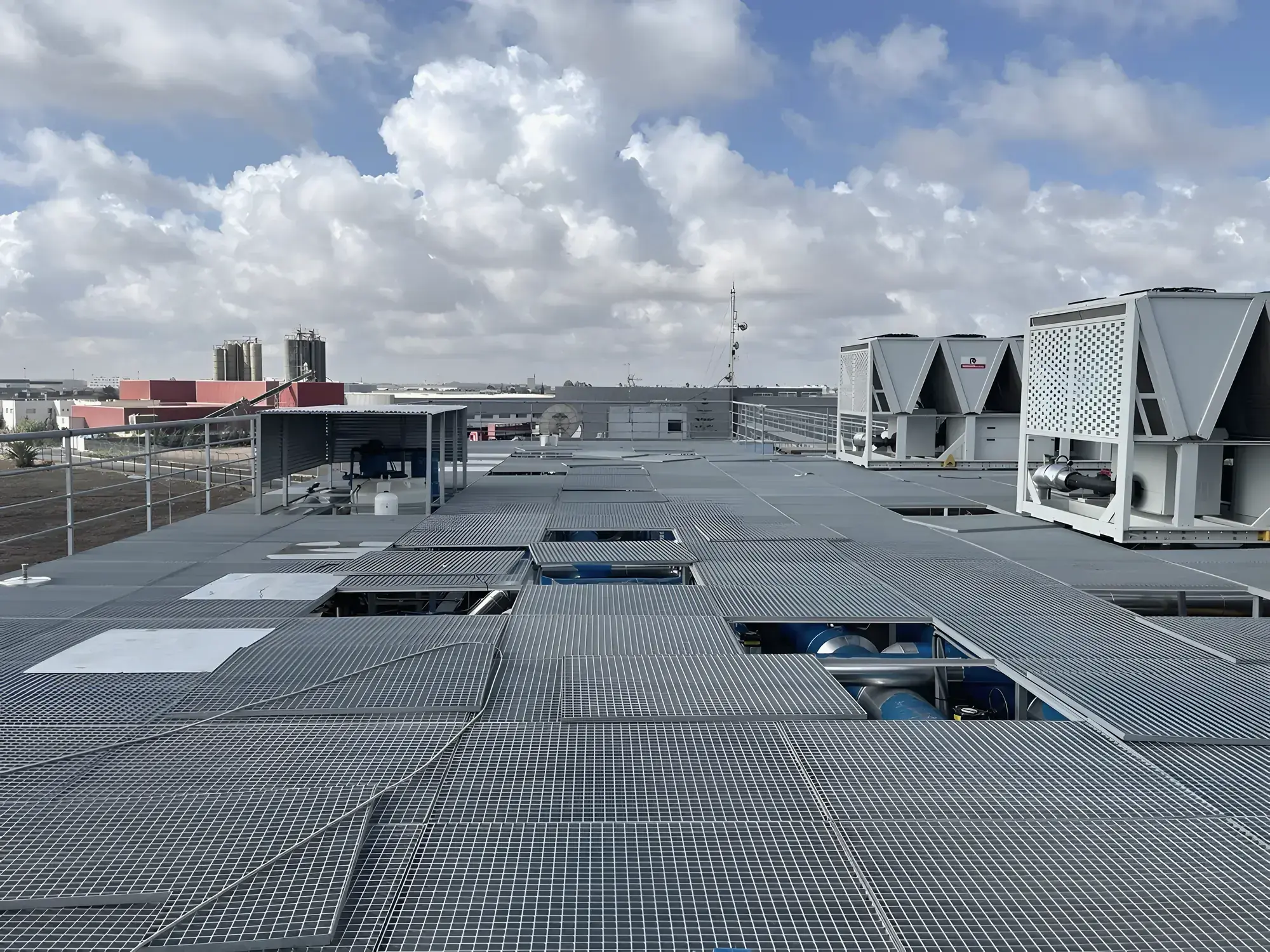 The roof deck seen across its steel grating, with the chillers at the far end and the industrial zone beyond under a cloud sky